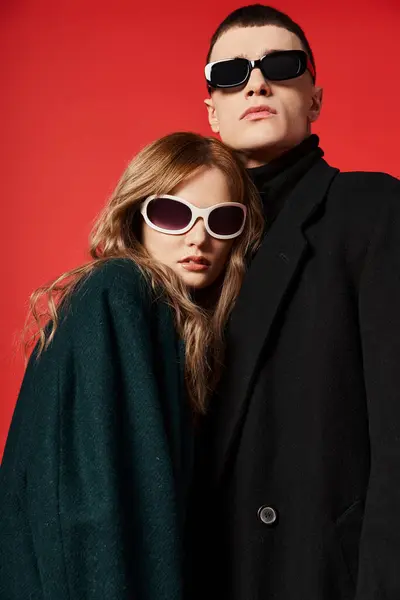 Beau jeune couple en manteaux élégants avec des lunettes de soleil posant ensemble sur fond rouge — Photo de stock