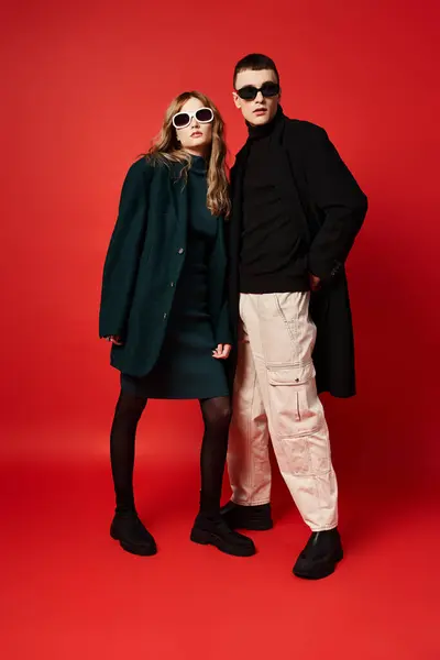 Bonito jovem casal em casacos elegantes com óculos de sol posando juntos no fundo vermelho — Fotografia de Stock