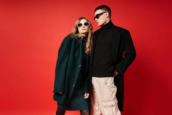 Gut aussehendes junges Paar in stylischen Mänteln mit Sonnenbrille posiert gemeinsam vor rotem Hintergrund — Stockfoto