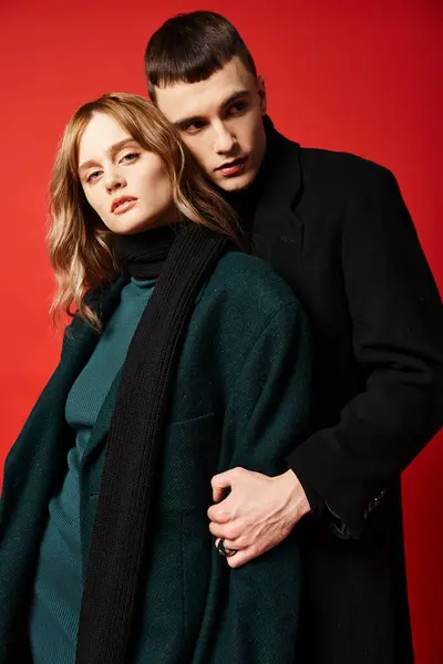 Attraktive Frau im Mantel blickt in die Kamera und posiert liebevoll mit ihrem Freund vor rotem Hintergrund — Stockfoto