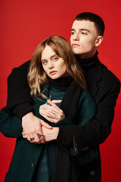 Appealing woman in coat looking at camera and posing lovingly with her boyfriend on red backdrop — Stock Photo