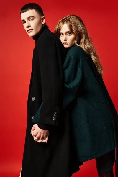 Очаровательная любящая пара в изысканных шикарных пальто, смотрящая на камеру на красном ярком фоне — стоковое фото