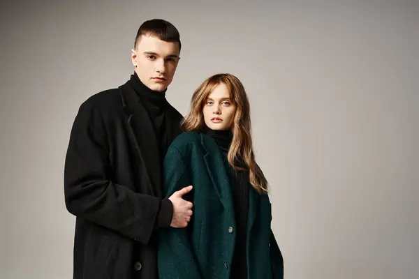 Модная пара в стильных пальто позирует вместе на сером фоне и смотрит в камеру — стоковое фото