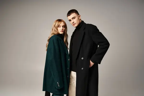 Модная пара в стильных пальто позирует вместе на сером фоне и смотрит в камеру — стоковое фото