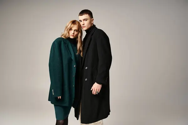 Сложная пара в стильных пальто позируют вместе на сером фоне и смотрят в камеру — стоковое фото