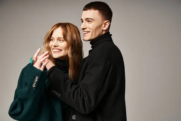 Freudig ansprechendes Paar in eleganten Mänteln, glücklich lächelnd und mit qualitativ hochwertiger Zeit auf grauem Hintergrund — Stockfoto
