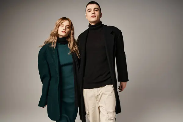 Appealing trendy boyfriend and girlfriend in coats posing and looking at camera on gray backdrop — Stock Photo
