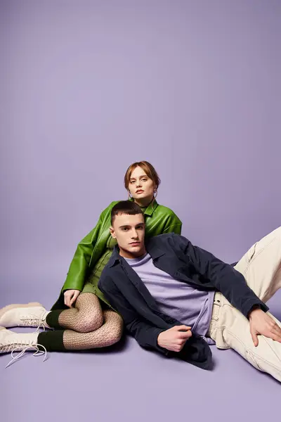 Good looking couple in vibrant attires sitting on floor and looking at camera on purple backdrop — Stock Photo