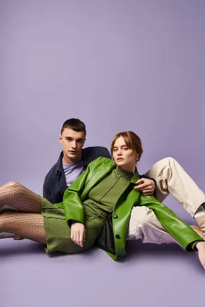 Sophisticated couple in vibrant attires sitting on floor and looking at camera on purple backdrop — Stock Photo