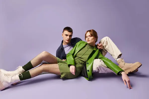 Модная пара в ярких нарядах, сидящая на полу и смотрящая на камеру на фиолетовом фоне — стоковое фото