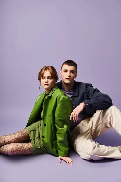 Модная пара в ярких нарядах, сидящая на полу и смотрящая на камеру на фиолетовом фоне — стоковое фото