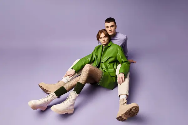Привлекательная пара в ярких одеждах, сидящих на полу и смотрящих на камеру на фиолетовом фоне — стоковое фото