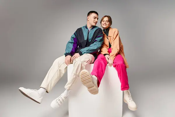 Cheerful woman in fashionable bomber looking at camera next to her handsome boyfriend on white cube — Stock Photo