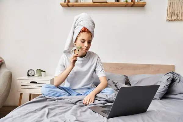 Séduisante personne queer aux cheveux rouges en vêtements de maison en utilisant un rouleau facial tout en se relaxant sur le lit avec un ordinateur portable — Photo de stock