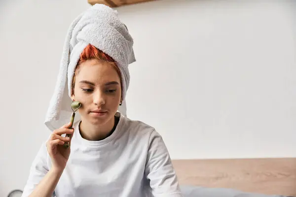 Hermosa persona joven extravagante en ropa de casa cómoda con toalla de pelo usando rodillo facial en casa - foto de stock