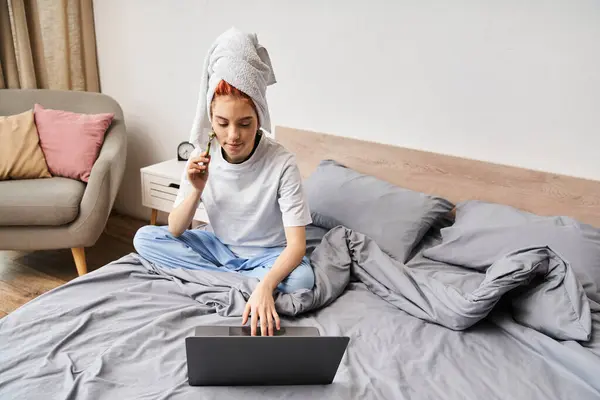 Séduisante personne queer aux cheveux rouges en vêtements de maison en utilisant un rouleau facial tout en se relaxant sur le lit avec un ordinateur portable — Photo de stock