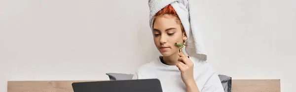 Atraente queer pessoa em homewear usando rolo rosto enquanto relaxa em sua cama com laptop, banner — Fotografia de Stock