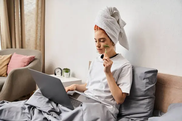 Atractivo pelirrojo queer persona en ropa de casa utilizando rodillo facial mientras se relaja en la cama con el ordenador portátil - foto de stock