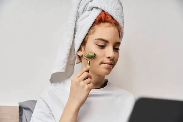 Atraente de cabelos vermelhos queer pessoa em casa usando rolo de rosto enquanto relaxa na cama com laptop — Fotografia de Stock