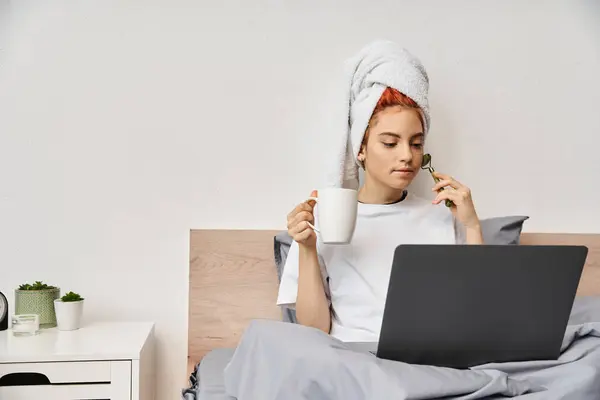 Belle personne queer avec serviette de cheveux en utilisant le rouleau de visage boire du thé tout en regardant des films à la télévision — Photo de stock