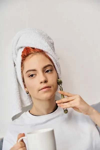 Attraente persona queer con asciugamano per capelli utilizzando rullo viso e tenendo la tazza di tè in mano mentre a casa — Foto stock