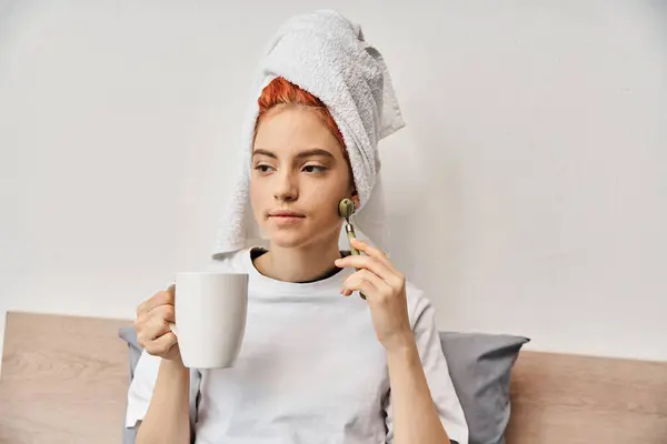 Atractiva persona queer con toalla de pelo usando rodillo facial y sosteniendo la taza de té en la mano mientras está en casa - foto de stock