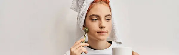 Attraente persona queer con asciugamano per capelli utilizzando rullo viso e tenendo tazza di tè mentre a casa, banner — Foto stock