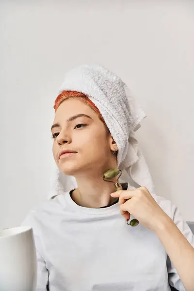 Attraente persona queer con asciugamano per capelli utilizzando rullo viso e tenendo la tazza di tè in mano mentre a casa — Foto stock