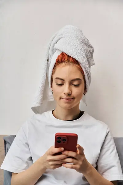 Joven persona extravagante en ropa de casa con toalla de pelo mirando su teléfono inteligente mientras se relaja en la cama - foto de stock