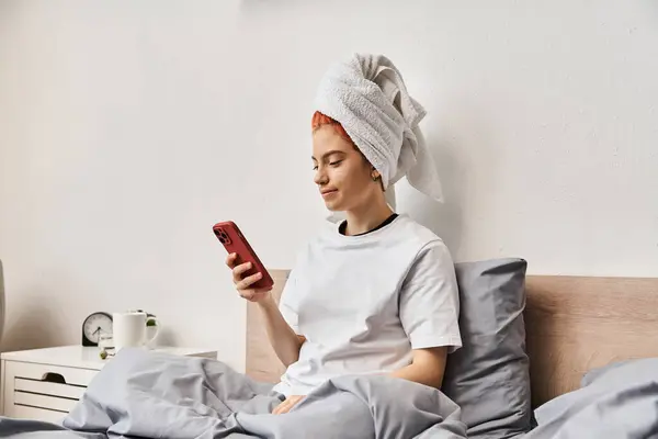 Молода екстравагантна людина в домашньому одязі з рушником, дивлячись на її смартфон, відпочиваючи в ліжку — стокове фото