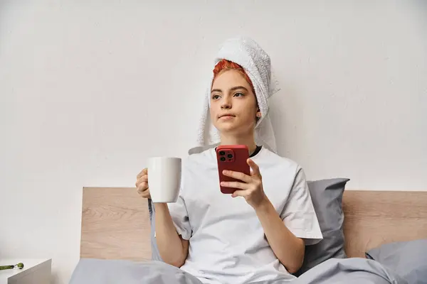 Positivo relaxante queer pessoa em casa com toalha de cabelo usando telefone e beber chá na cama — Fotografia de Stock