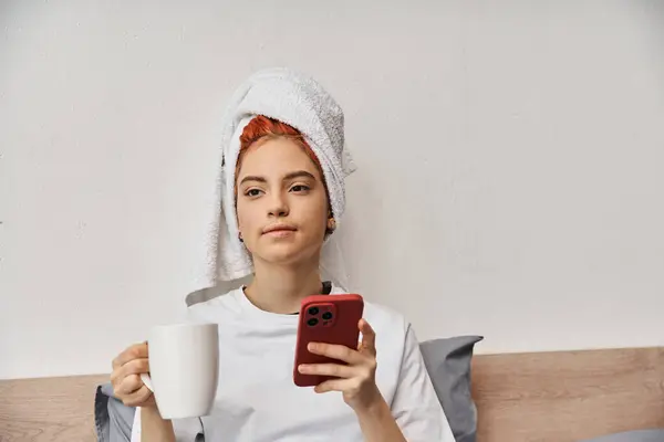 Positivo relaxante queer pessoa em casa com toalha de cabelo usando telefone e beber chá na cama — Fotografia de Stock