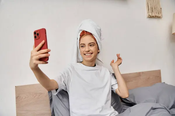 Alegre hermosa persona queer con toalla de pelo en la ropa de casa tomar selfies mientras se relaja en la cama - foto de stock