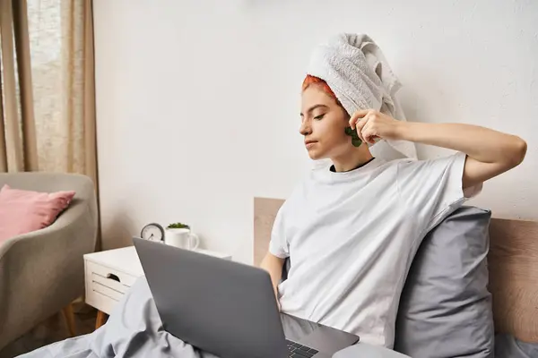 Alegre muito queer pessoa com toalha de cabelo assistindo filmes no laptop e usando gua sha enquanto na cama — Fotografia de Stock