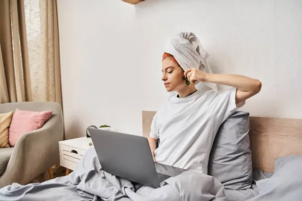 Gioiosa bella persona queer con asciugamano per capelli guardando film sul computer portatile e utilizzando gua sha mentre a letto — Foto stock