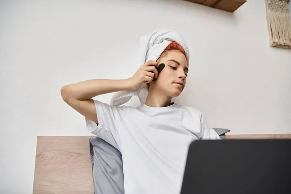 Alegre muito queer pessoa com toalha de cabelo assistindo filmes no laptop e usando gua sha enquanto na cama — Fotografia de Stock