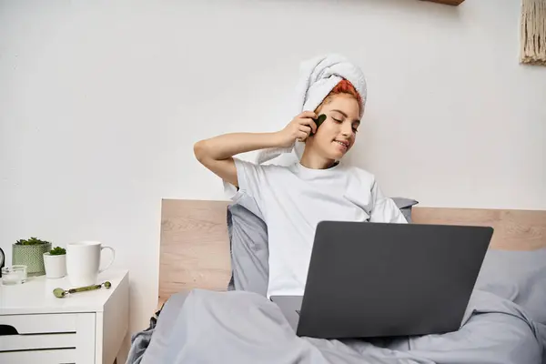 Alegre muito queer pessoa com toalha de cabelo assistindo filmes no laptop e usando gua sha na cama — Fotografia de Stock