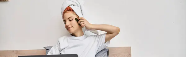 Joyeux jolie personne queer avec serviette de cheveux regarder des films sur ordinateur portable et en utilisant gua sha au lit, bannière — Photo de stock