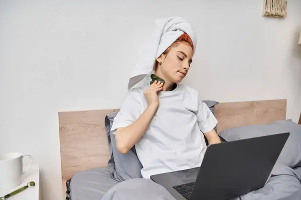Allegro bella persona queer con asciugamano per capelli guardando film sul computer portatile e utilizzando gua sha a letto — Foto stock
