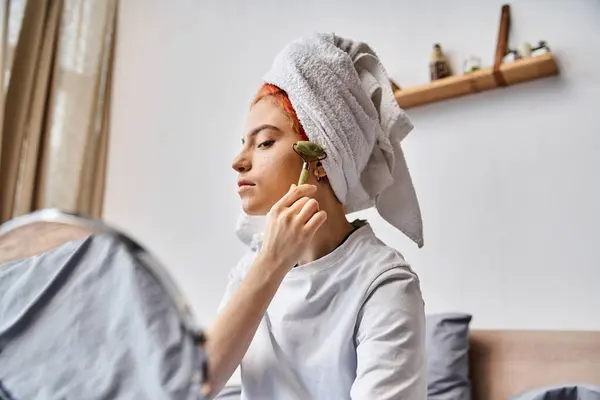 Good looking queer person in cozy homewear using face roller in front of mirror as morning routine — Stock Photo