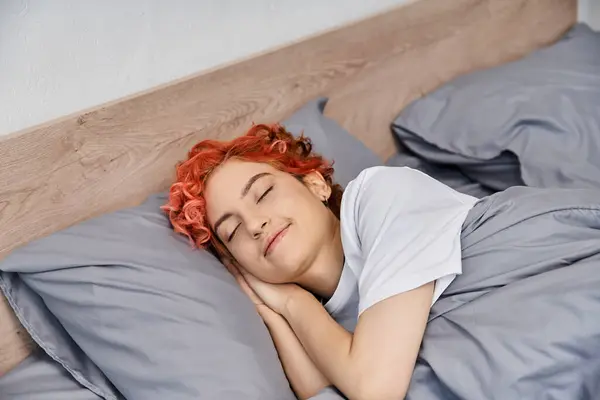 Bien parecido pelirrojo queer persona en acogedora ropa de casa siesta en su cama en casa, tiempo libre - foto de stock