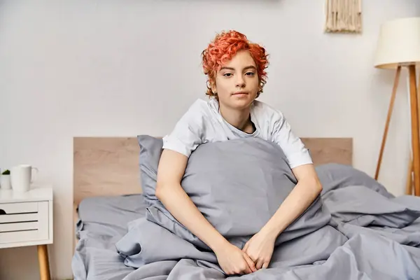 Sonolento extravagante queer pessoa em homewear com cabelo vermelho sentado na cama e olhando para a câmera — Fotografia de Stock