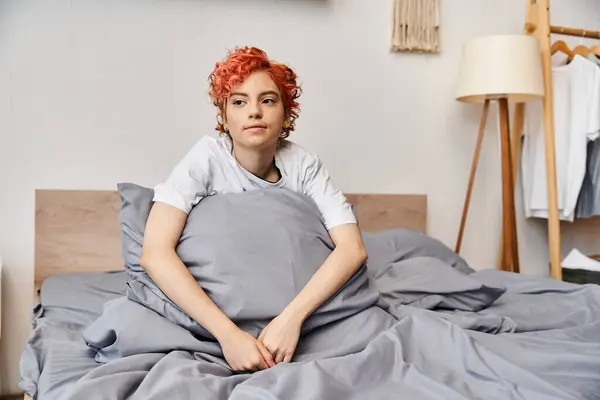 Entspannte rothaarige queere Person in Hauskleidung, die zu Hause auf ihrem Bett sitzt und wegschaut, Freizeit — Stockfoto