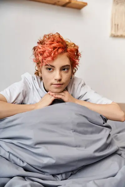 Relajante pelirroja queer persona en ropa de casa sentado en su cama en casa y mirando hacia otro lado, ocio - foto de stock