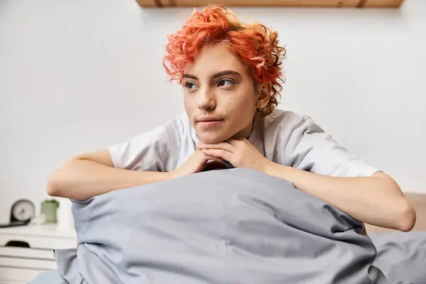 Rilassante persona dai capelli rossi queer in biancheria da casa seduto sul suo letto a casa e guardando lontano, tempo libero — Foto stock