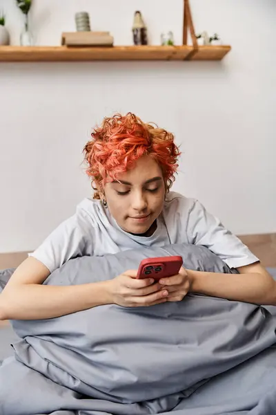 Soñoliento hermosa queer persona en ropa de casa con el pelo rojo sentado en la cama y el uso de su teléfono inteligente - foto de stock