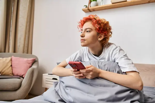 Сонный привлекательный странный человек в домашней одежде с рыжими волосами, сидящий на кровати и использующий ее смартфон — стоковое фото