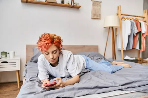 Bela pessoa queer relaxante em roupa de casa com cabelo vermelho deitado na cama e usando seu smartphone — Fotografia de Stock