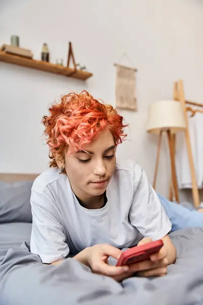 Hermosa persona queer relajante en ropa de casa con el pelo rojo acostado en la cama y el uso de su teléfono inteligente - foto de stock