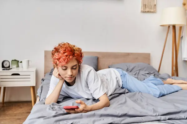 Hermosa persona queer relajante en ropa de casa con el pelo rojo acostado en la cama y el uso de su teléfono inteligente - foto de stock